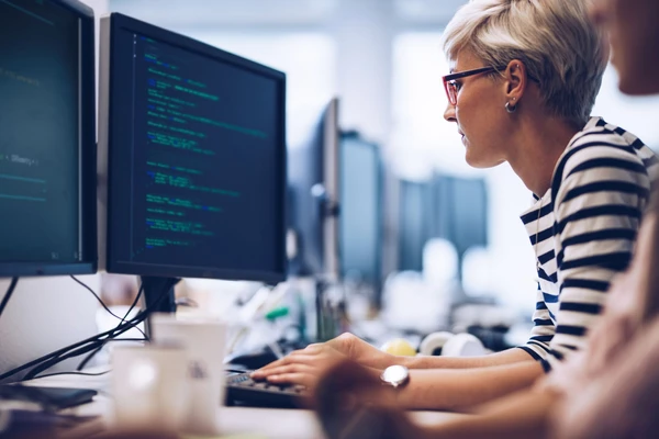 short-haired blonde woman with re glasses looks at code on desktop monitor